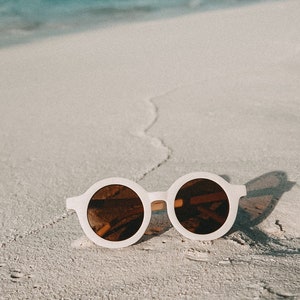 May include: White round sunglasses with brown lenses on a white sandy beach.