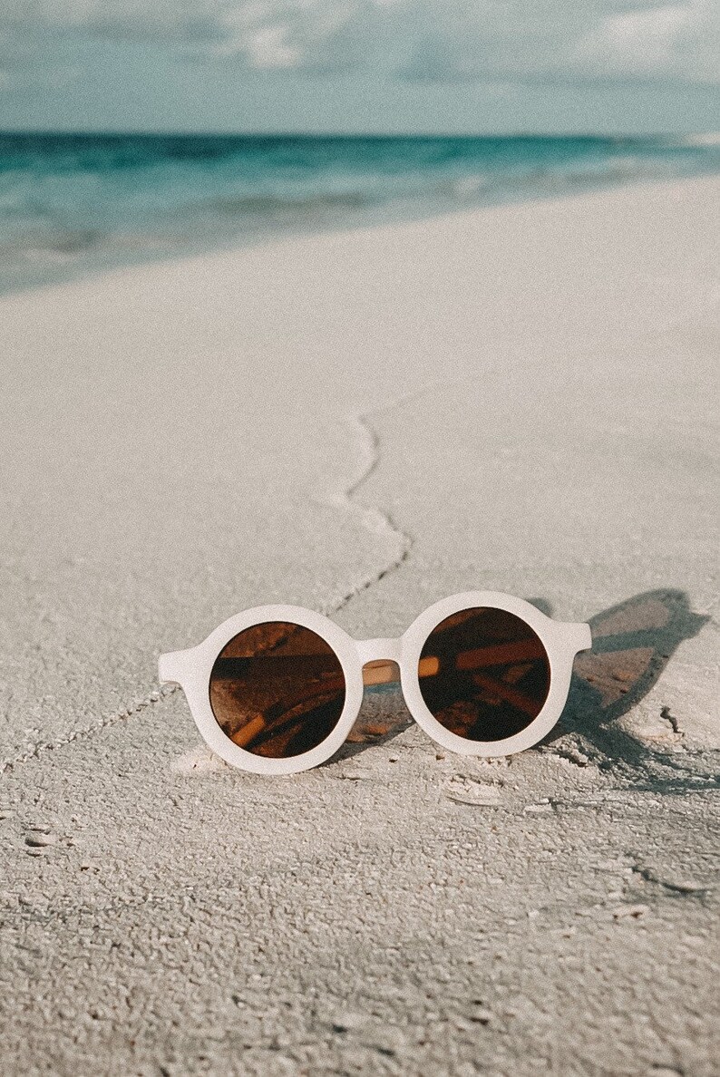 May include: White round sunglasses with brown lenses on a white sandy beach.