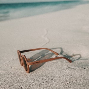 May include: Brown round sunglasses with the text "Petit Panda" on the frame, lying on a white sandy beach.