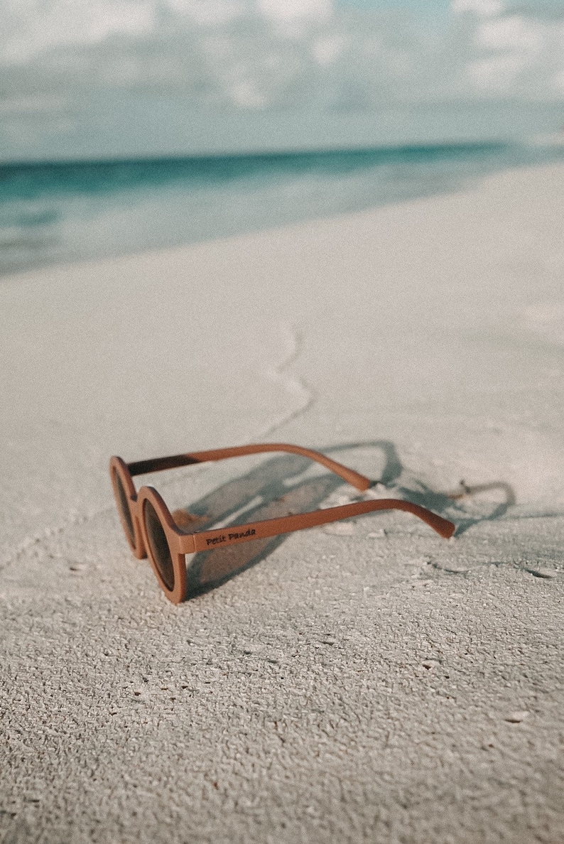 May include: Brown round sunglasses with the text "Petit Panda" on the frame, lying on a white sandy beach.