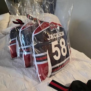 Puede incluir: Tres camisetas deportivas negras y rojas en embalaje de plástico transparente. Cada camiseta tiene el nombre "Jackson" y el número "58" en blanco. Las camisetas tienen rayas blancas y detalles rojos.