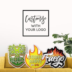 May include: Three colorful logo-shaped stickers on a yellow couch. The stickers feature the text "EDEN", "FRUIT RIOT! got spicy", and "Fuego". Above, a black-framed sign reads "Customize with your logo".