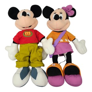 May include: A pair of plush Mickey Mouse and Minnie Mouse toys. Mickey is wearing a red shirt with a green "M Mickey" logo, yellow pants, and blue shoes. Minnie is wearing an orange shirt with white stars, a purple skirt, and purple shoes.