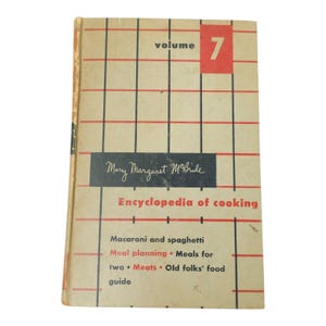Puede incluir: Un libro de tapa dura vintage titulado Mary Margaret McBride Encyclopedia of cooking volume 7. La portada presenta un fondo beige con un patrón de cuadrícula rojo y negro, texto en negro y detalles en rojo que enumeran temas culinarios.