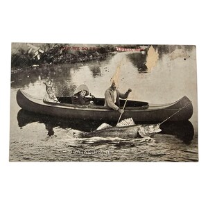 May include: A black and white vintage postcard showing three people in a canoe with a large fish caught on a fishing line. The text on the postcard reads "How We Do This - Tupelo, Miss. -  Fishing Here Is Fun."