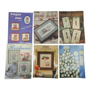 May include: A selection of vintage cross-stitch pattern books. Titles include "Pitiful Pals," "Home Again," "Garden Medley," "Heirlooms," and a Coca-Cola themed design. The books feature framed designs and illustrations.
