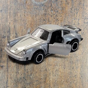 May include: A silver die-cast toy sports car with a rear spoiler, black wheels, and an open driver-side door. The vehicle features a sloped hood, round headlights, and a detailed interior visible through the open door and windows.