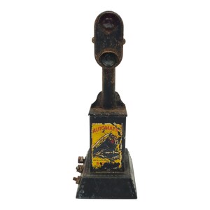 May include: Vintage black metal toy train signal with a red and green light. The signal has a rectangular base and a box with the word "AUTOMATIC" on it. The signal is weathered and shows signs of age.