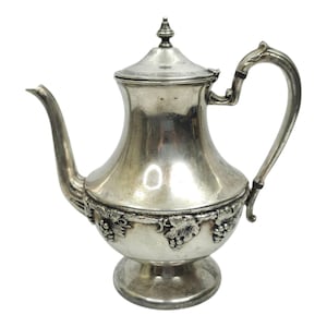 Puede incluir: Una tetera de metal plateado con un pico curvo, una tapa con un remate puntiagudo y un asa ornamentada. La base del cuerpo está decorada con un patrón en relieve de racimos de uvas y hojas de parra que rodea toda la circunferencia.
