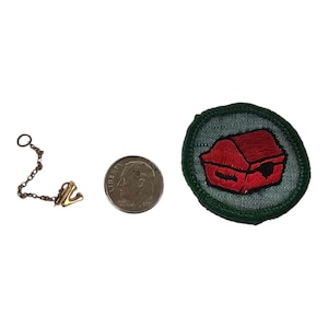 May include: A gold-toned metal charm shaped like the letter V attached to a small chain, a silver-colored US dime for scale, and a circular green embroidered patch featuring a red barn-like structure with black outlines and details.