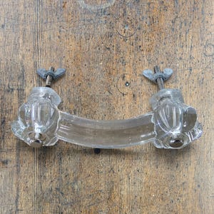 May include: A clear glass drawer pull with a curved handle and two fluted, knob-like ends. The hardware features two threaded metal bolts extending from the back, each secured with a silver-toned metal wing nut for mounting purposes.