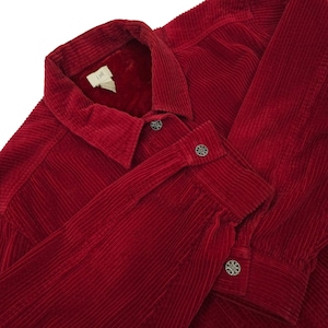 May include: A close-up of a red corduroy shirt with a collar and long sleeves. The shirt has decorative buttons on the cuffs and front. The fabric has a ribbed texture. The shirt is a deep shade of red.