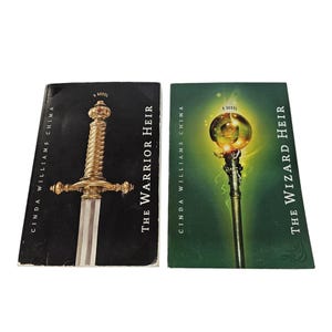 May include: Two paperback books by Cinda Williams Chima. The left book is black with a gold sword illustration and the title The Warrior Heir. The right book is green with a glowing orb staff illustration and the title The Wizard Heir.