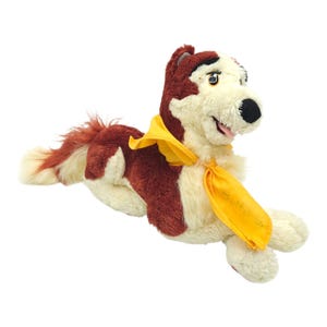 May include: A soft toy dog in a lying down pose. The toy is red and cream-coloured with a yellow scarf. The dog has a black nose and a smiling expression. The scarf has the text "Balto" printed on it.