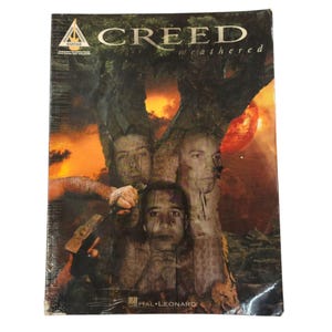 May include: A guitar music book titled "CREED weathered" featuring a band photo with a tree and a red orb. The cover has a dark, weathered aesthetic with the Hal Leonard logo at the bottom.