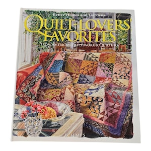 May include: A softcover book titled Better Homes and Gardens Quilt-Lovers' Favorites Volume 5. The cover features a colorful patchwork quilt draped over a red sofa, with a glass bowl of flowers in the foreground and a sunroom in the background.