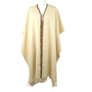 May include: A beige poncho with a V-neck and a dark brown stitched trim down the front. The poncho has a fringed hemline. The garment is displayed on a mannequin against a white background.