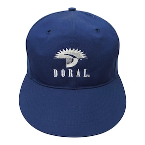 May include: A solid navy blue baseball cap featuring an embroidered white logo on the front panel. The logo consists of a stylized sunburst graphic above the text "DORAL" in a serif font. The cap has a structured crown and a curved brim.