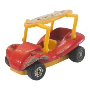 May include: A vintage toy car in red with yellow roll bars and a surfboard on top. The toy car has black wheels and a yellow interior. The surfboard has faded text. The toy car is a miniature version of a dune buggy.