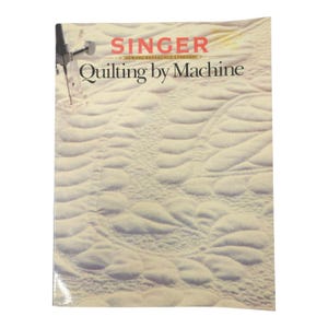 May include: A Singer Sewing Reference Library book titled "Quilting by Machine." The cover shows a close-up of quilted fabric with a sewing machine needle and the Singer logo in red. The background is a light beige colour.