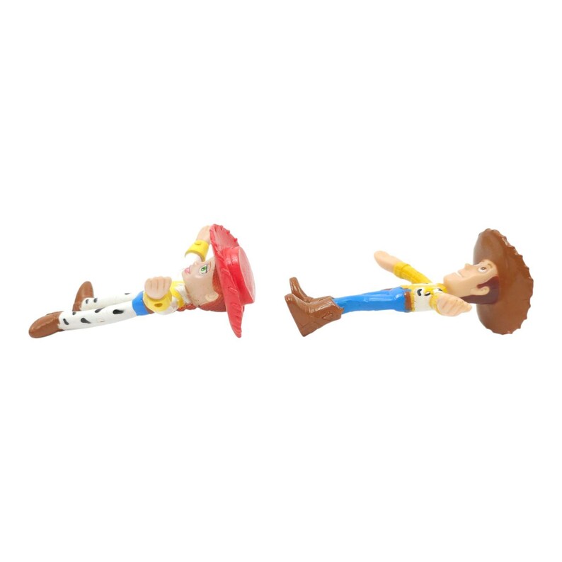 May include: Two Toy Story character figurines, Jessie and Woody, are lying down. Jessie wears a red hat, yellow shirt, and cow-print trousers. Woody wears a brown hat, yellow shirt, blue trousers, and brown boots. Both are on a white background.
