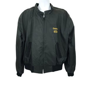 Puede incluir: Una chaqueta negra con cremallera con un logotipo de Panasonic bordado en oro en el pecho izquierdo. La chaqueta tiene un cuello alto y puños y cintura de canalé.