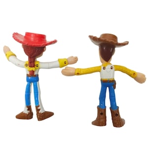 May include: Two Toy Story character figurines, Jessie and Woody, stand side-by-side. Jessie wears a red hat, white shirt, blue trousers, and brown boots. Woody wears a brown hat, yellow shirt, blue trousers, and brown boots. Both have outstretched arms.
