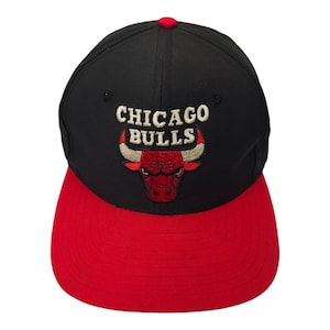 Puede incluir: Gorra de béisbol negra y roja con el logotipo de los Chicago Bulls bordado en la parte delantera. El logotipo presenta un toro rojo con cuernos negros y un contorno blanco. El texto "CHICAGO BULLS" está bordado en dorado sobre el toro.