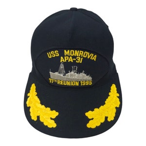 May include: A navy blue baseball cap with a yellow embroidered patch that reads "USS MONROVIA APA-31 11TH REUNION 1995". The patch also features a gray embroidered image of a ship. The cap has two yellow embroidered laurel wreaths on the bill.
