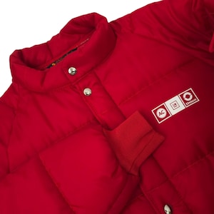 Peut inclure: Veste matelassée rouge vif avec un col montant et des boutons-pression argentés. La veste présente un poignet côtelé rouge et un écusson blanc avec les inscriptions "AC", "GM" et "Delco".