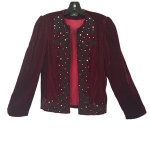 May include: A burgundy velvet jacket with long sleeves and a black embroidered design. The open front jacket features a decorative pattern with silver-coloured embellishments. The jacket is hanging on a black hanger.