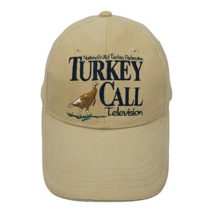 May include: A tan baseball cap with the text "TURKEY CALL Television" embroidered in navy blue. The cap also features an embroidered image of a turkey. The text "National Wild Turkey Federation" is above the main text.