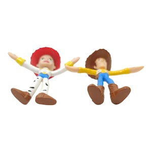 May include: Two small Toy Story character figurines: Jessie and Woody. Jessie has a red hat, white shirt, and blue trousers with black spots. Woody wears a brown hat, yellow shirt, and blue trousers.