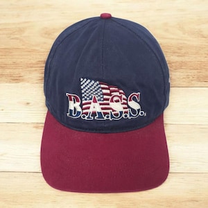May include: A navy blue baseball cap with a maroon brim. The cap features the word "BASS" embroidered in white, red, and blue, along with a stylized American flag design. The cap is made of a durable fabric.