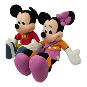 May include: A plush Mickey Mouse and Minnie Mouse toy set. Mickey is wearing a red shirt with the word "Mickey" on it. Minnie is wearing an orange shirt with a purple scarf and a pink bow.