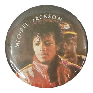 May include: A round button featuring a portrait of Michael Jackson. The button has a dark gray background with the words "MICHAEL JACKSON" printed in white. Jackson is depicted wearing a red jacket and looking towards the viewer.