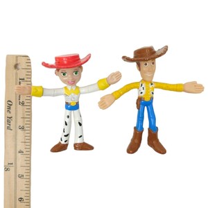 May include: Two articulated action figures of the characters Jessie and Woody from the animated film Toy Story. Jessie wears a red hat, white shirt, and cow-print trousers. Woody wears a brown hat, yellow shirt, and blue trousers. The figures are approximately 13 cm tall.