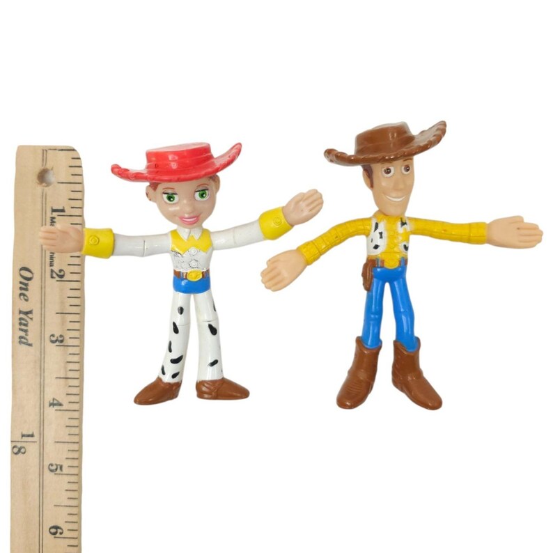 May include: Two articulated action figures of the characters Jessie and Woody from the animated film Toy Story. Jessie wears a red hat, white shirt, and cow-print trousers. Woody wears a brown hat, yellow shirt, and blue trousers. The figures are approximately 13 cm tall.
