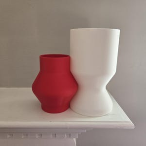 May include: Two ceramic vases, one red and one white, sit side-by-side on a white shelf. The red vase has a rounded shape, while the white vase is taller with a unique hourglass design. The background is a light gray wall.