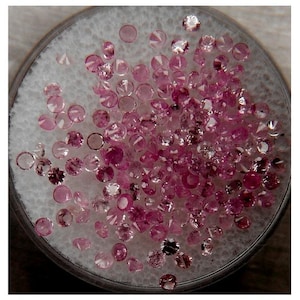 May include: A close-up of a collection of pink gemstones in a white foam-lined dish. The gemstones are various sizes and shapes, with some having a more intense pink hue than others.