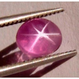 Puede incluir: Una piedra preciosa de zafiro estrellado rosa con una estrella de seis puntas en el centro. La piedra preciosa está sujeta por unas pinzas.