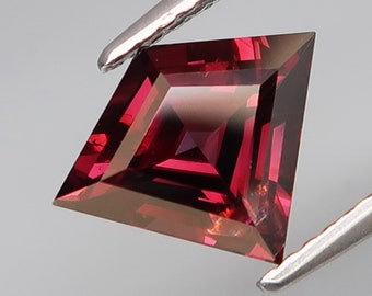 May include: A deep red gemstone with a square shape and a faceted cut. The gemstone is held in tweezers.