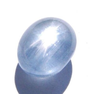 Puede incluir: Gema ovalada de color azul claro con efecto de estrella. La gema pulida refleja la luz, creando un patrón de estrella. Se proyecta una sombra debajo. El efecto de estrella se debe a inclusiones internas.