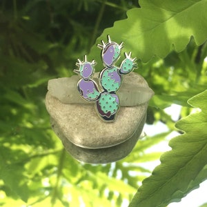 Peut inclure: Épingle en émail en forme de cactus avec un contour argenté. L'épingle présente un motif avec des nuances de violet et de sarcelle, avec de petits points verts. L'épingle repose sur une petite pierre de couleur claire.