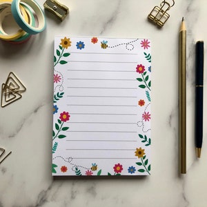 May include: A white notepad with a floral border and bees. The border features pink, yellow, blue, and green flowers. The notepad has lined paper.