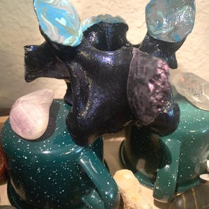 May include: A black, iridescent, ceramic sculpture of a stylized animal with two blue and white marbled wings. The sculpture is sitting on two green mugs with white speckles.
