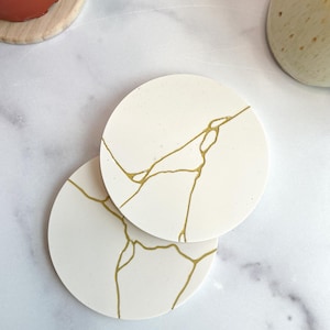 May include: Two circular white coasters featuring a gold-colored vein pattern that mimics the appearance of kintsugi. The coasters have a smooth, matte finish and are arranged in an overlapping position, showcasing the intricate gold lines.