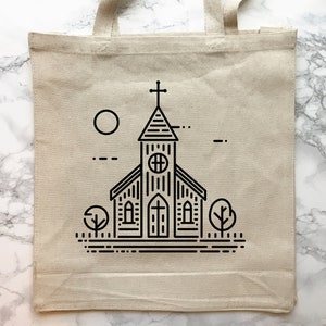 May include: A beige canvas tote bag featuring a black line art illustration of a church building with a steeple, a cross, a circular window, and arched doors. The design includes a sun, two stylized trees, and horizontal ground lines.