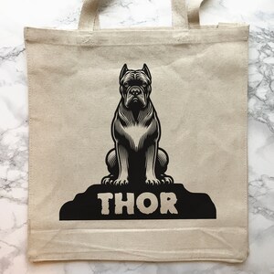 May include: A beige tote bag with black handles and a black and white illustration of a dog sitting on a rock with the word "THOR" printed below it.