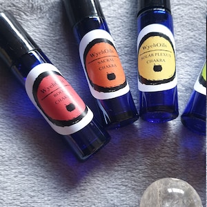 Puede incluir: Cuatro botellas de vidrio azul con rodillo y etiquetas para los diferentes chakras. Las etiquetas son naranja, roja, amarilla y rosa con texto negro. El texto en las etiquetas dice "WychOils" y el nombre del chakra. Las botellas están dispuestas en semicírculo sobre una superficie gris y peluda. Un cristal de cuarzo transparente con una piedra de amatista púrpura está en el centro de la imagen.